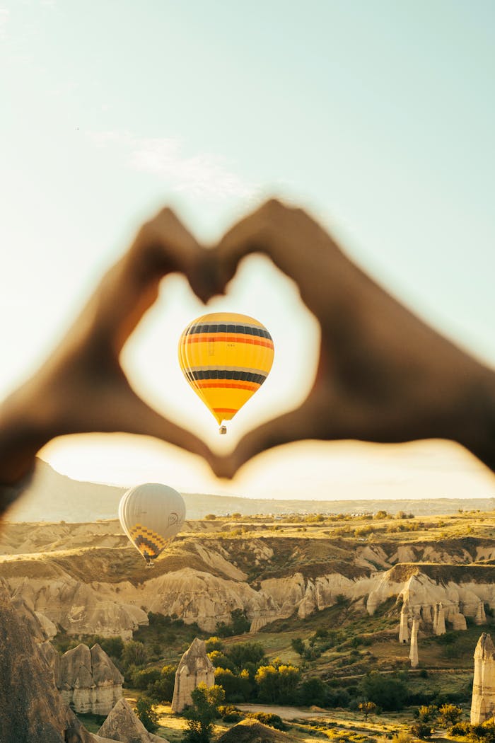 contact-img Romantic view of hot air balloons over Cappadocia at sunrise, framed by hands forming a heart.