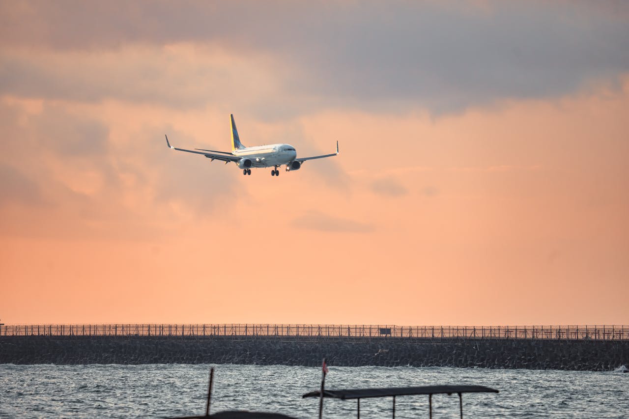 hero-img-01 Airplane descending over ocean towards Bali Airport with sunset sky backdrop.